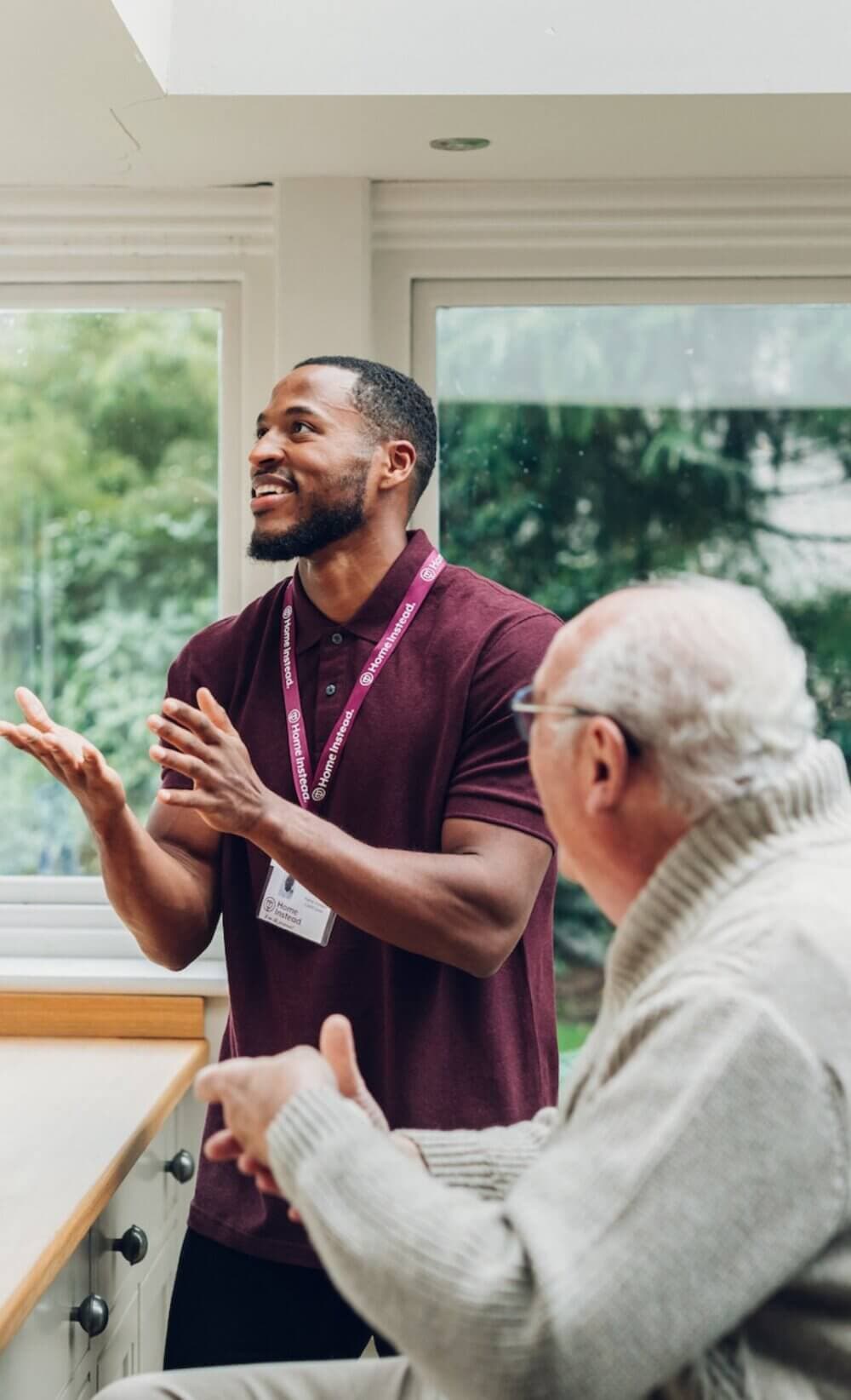 A man wearing a lanyard gestures while talking to an older man with glasses, seated at a table in a bright room. - Home Instead