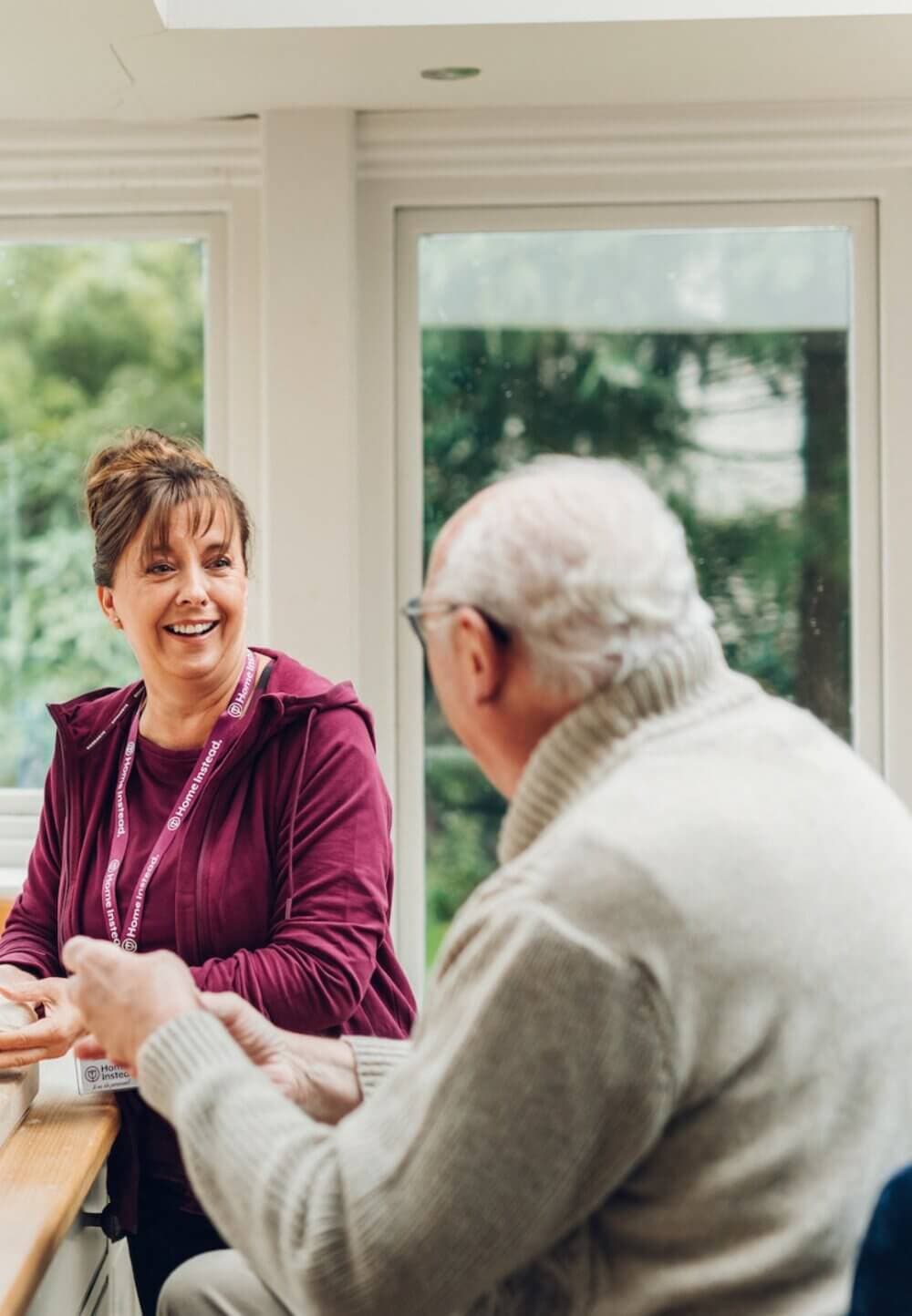A woman with a lanyard smiles while sitting at a table with an elderly man in a cozy, well-lit room. - Home Instead