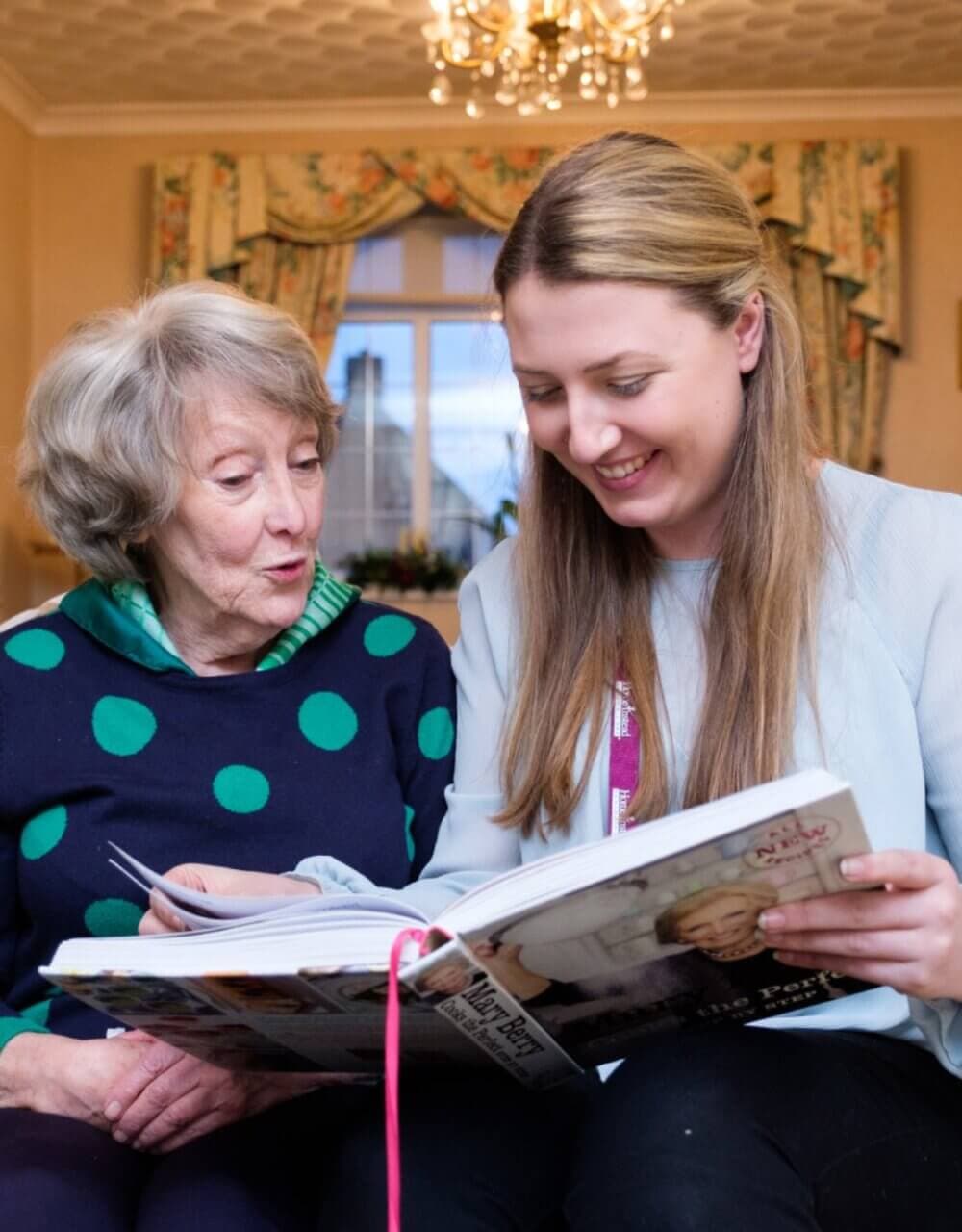 An elderly woman and a young woman smile while looking at a book together in a cozy room with a window and curtains. - Home Instead Poole