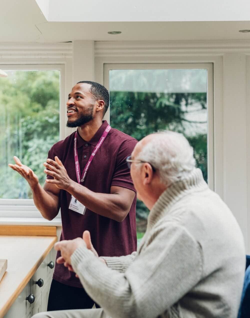 A young man and an elderly man have a conversation in a bright room with large windows. - Home Instead