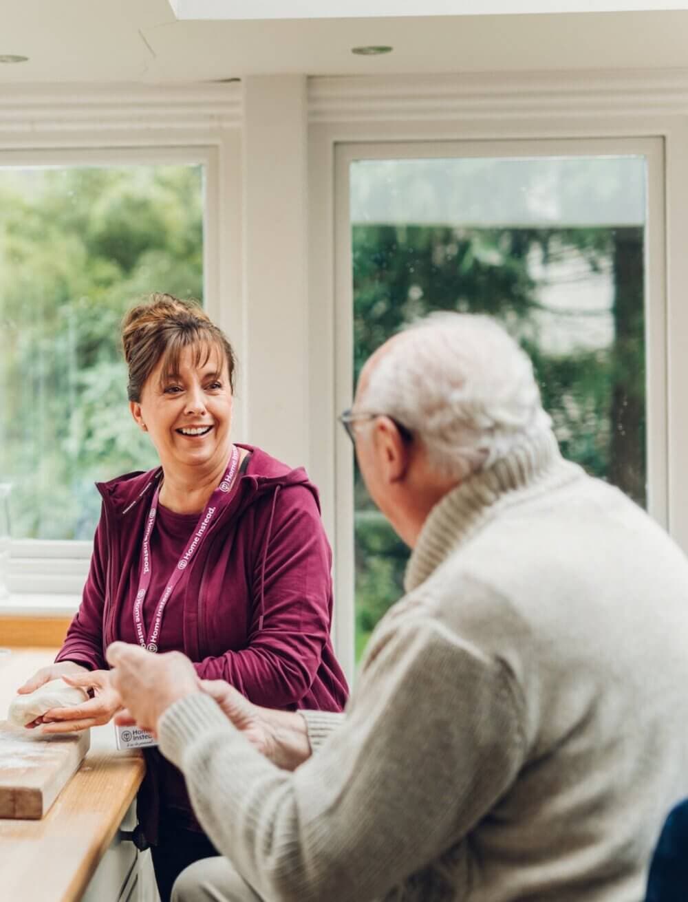 A woman in a purple jacket chats with an elderly man in a white sweater by a window. Both are smiling. - Home Instead