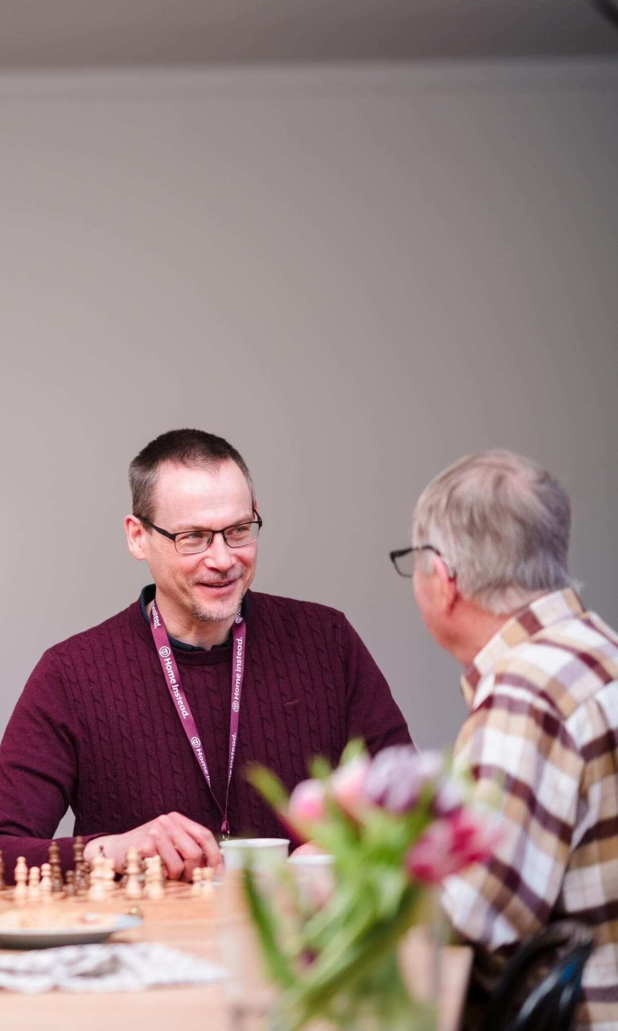 Two men, one in a maroon sweater and the other in a plaid shirt, conversing cheerfully over a chessboard. - Home Instead