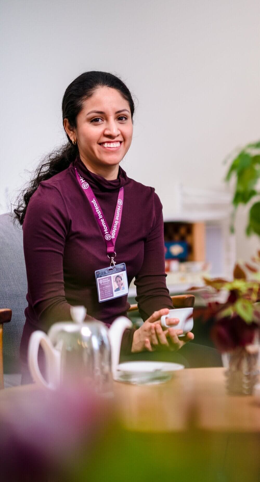 A smiling woman with dark hair sits holding a cup, wearing a burgundy top and an ID badge on a lanyard around her neck. - Home Instead Poole
