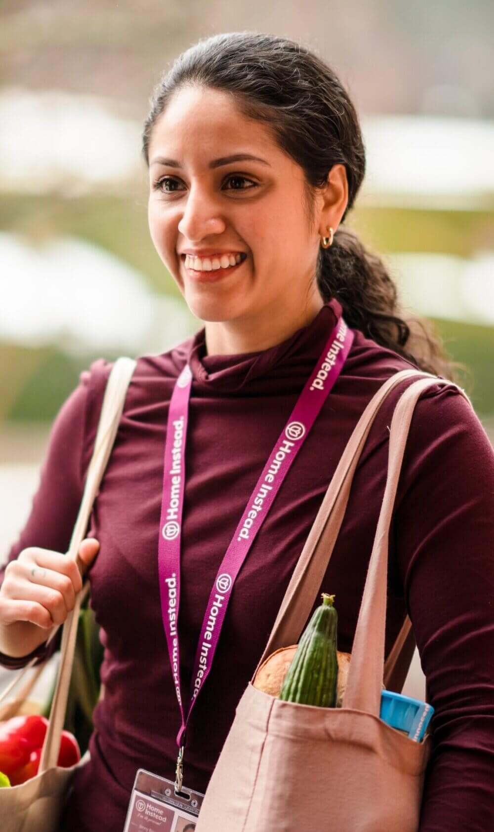 Smiling woman with a Home Instead lanyard, holding grocery bags filled with a zucchini and other items. - Home Instead