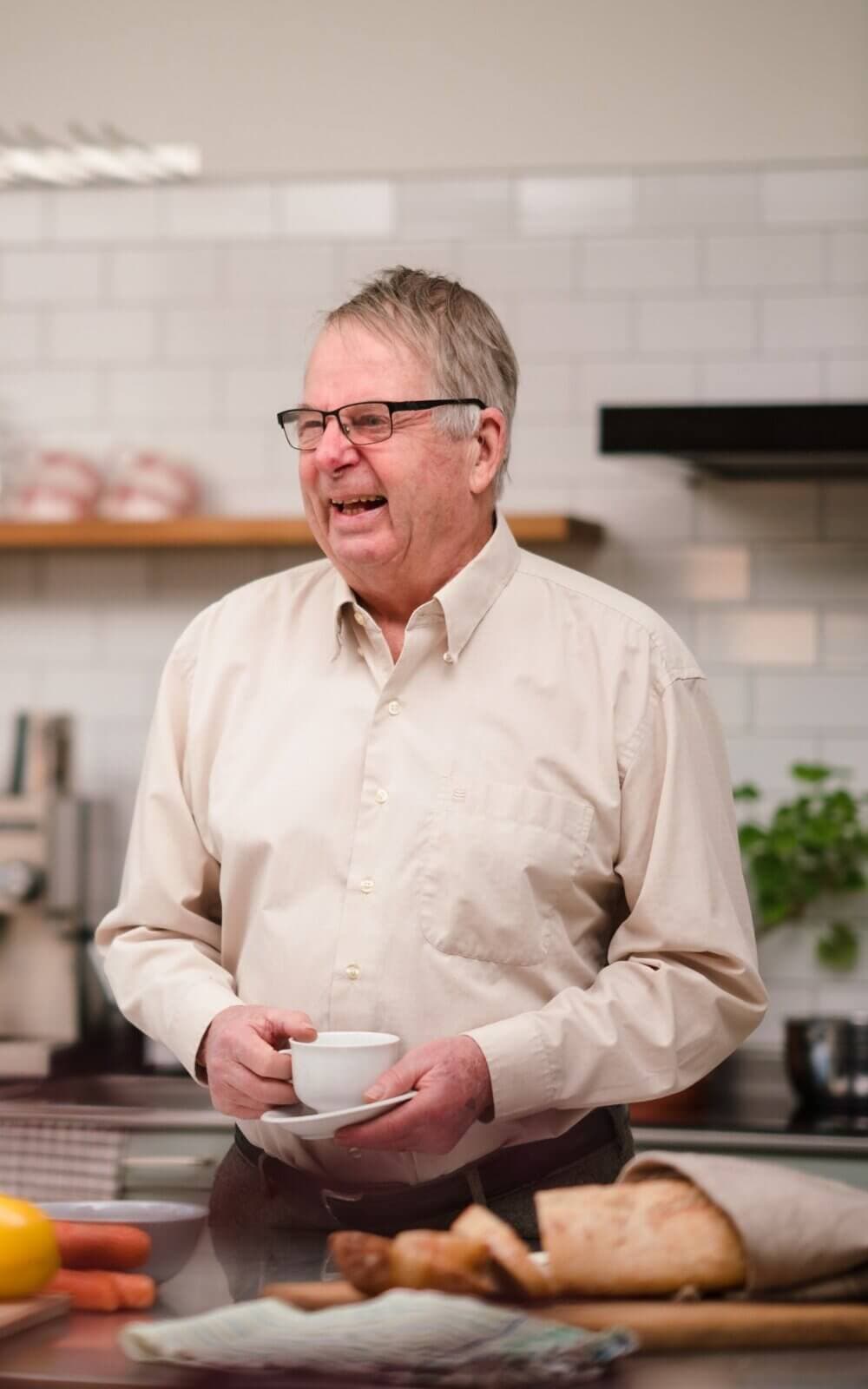 Smiling elderly man in a white shirt holding a teacup in a kitchen with bread and vegetables on the counter. - Home Instead Bournemouth & Christchurch