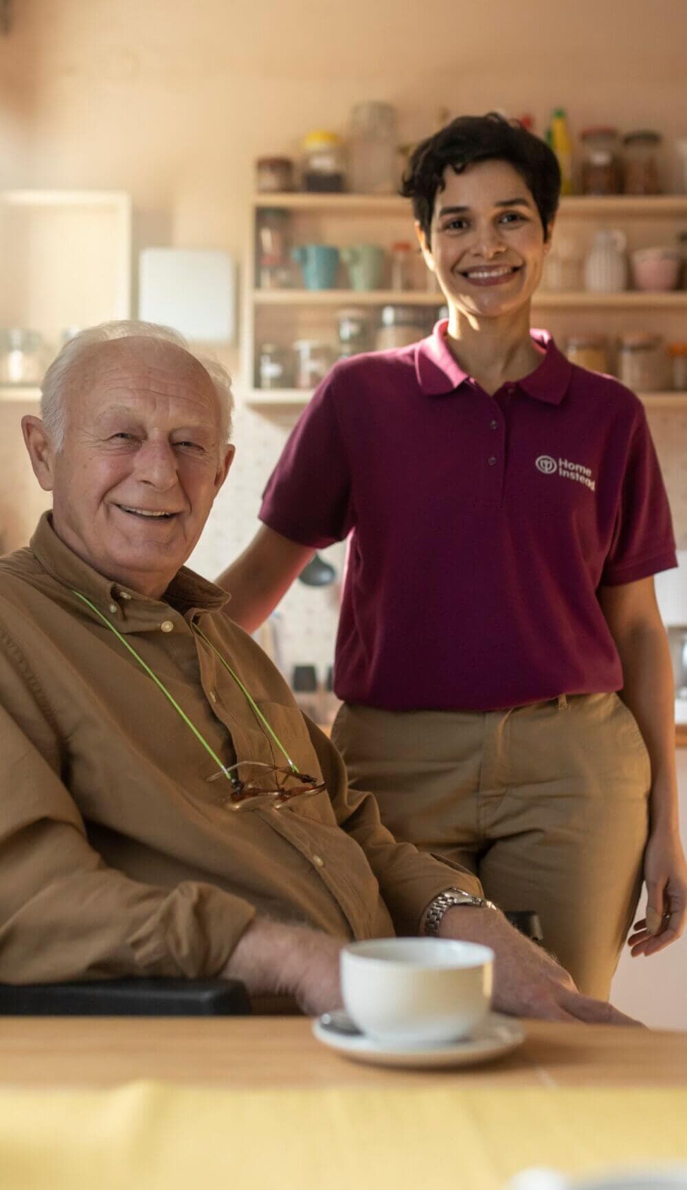 An elderly man and a caregiver smiling in a kitchen, with a coffee cup on the table in front of them. - Home Instead