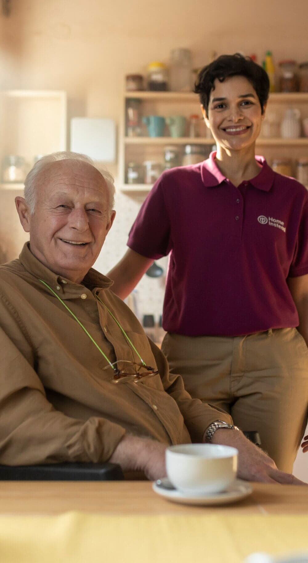 An elderly man smiles while seated at a table with a cup, and a caregiver stands beside him in a kitchen-like setting. - Home Instead