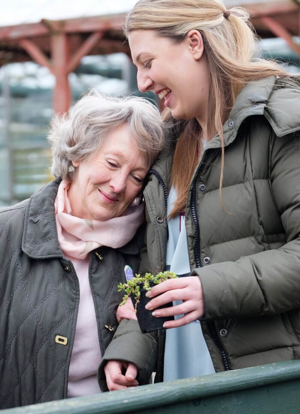 An older woman and a younger woman share a happy moment holding a small potted plant outdoors. - Home Instead Southampton