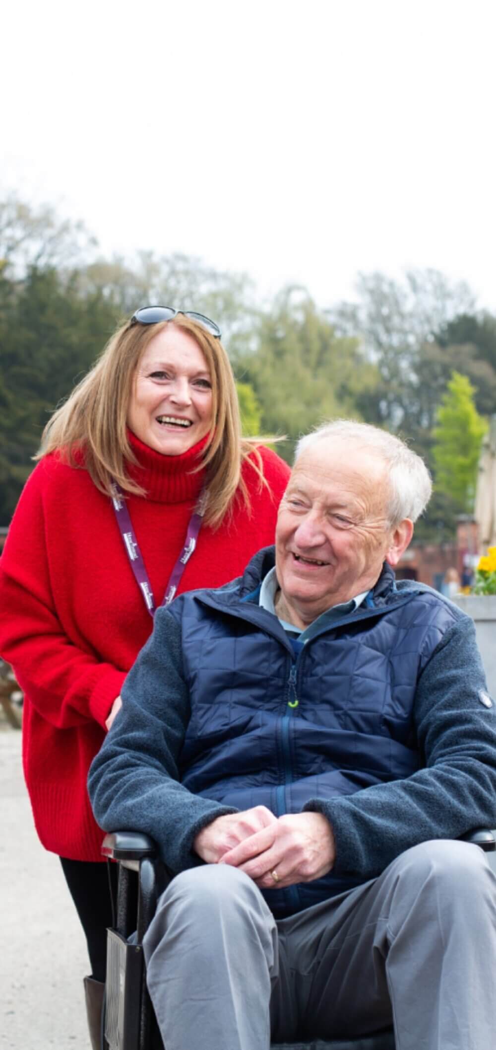 A smiling woman in a red sweater stands behind an older man in a wheelchair outdoors on a bright day. - Home Instead