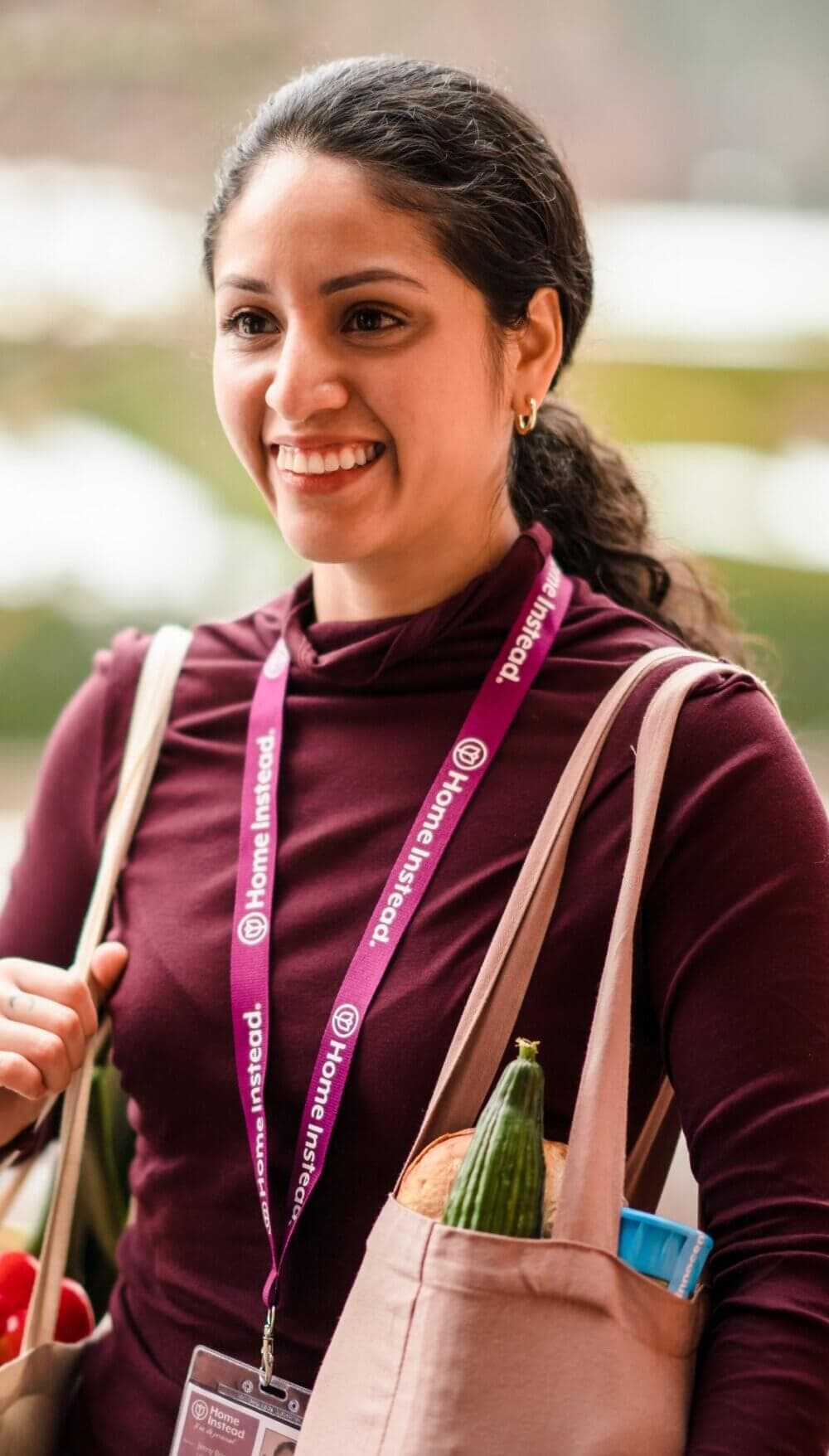 Woman smiling, wearing a lanyard, and carrying a grocery bag with produce and other items in it. - Home Instead