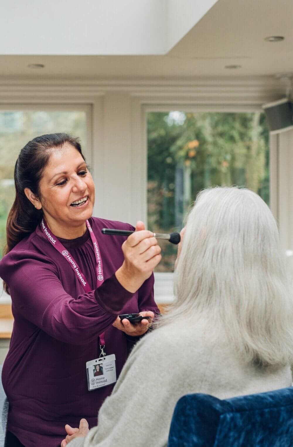 A Care Professional in a maroon top applies makeup to an elderly woman with long white hair seated in a chair. - Home Instead Bournemouth & Christchurch