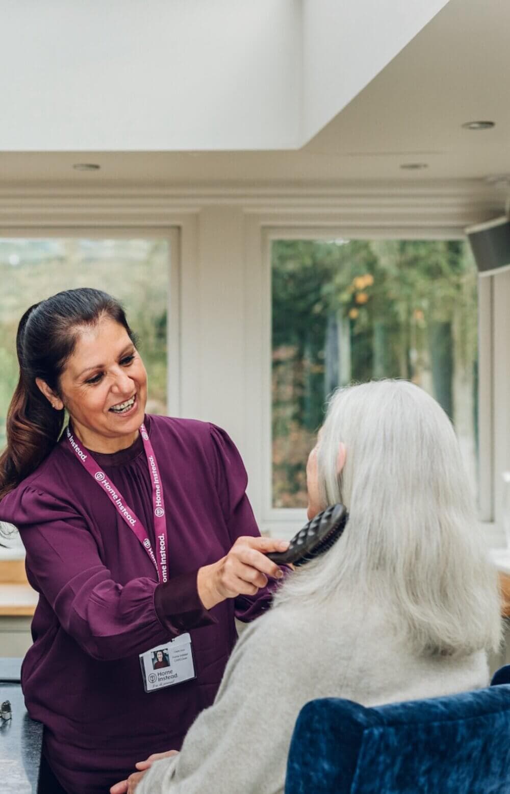 A Care Professional in a purple shirt brushes the hair of an elderly person with long white hair in a sunny room. - Home Instead Poole