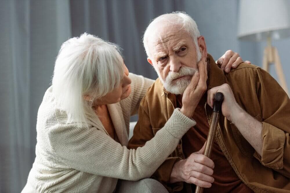 An elderly woman comforts a sad elderly man holding a cane, gently touching his face with concern. - Home Instead