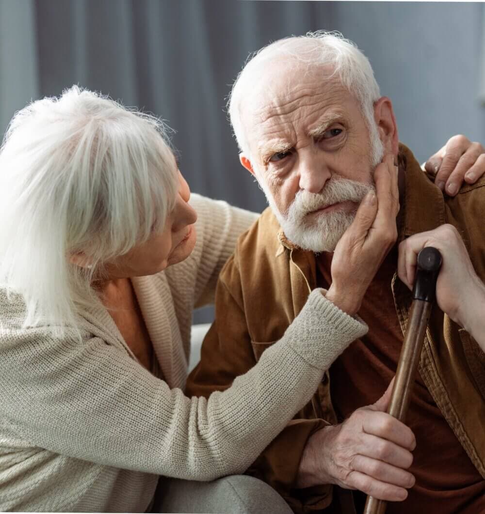 An elderly woman gently holds the face of an elderly man with a concerned expression, while he holds a walking stick. - Home Instead