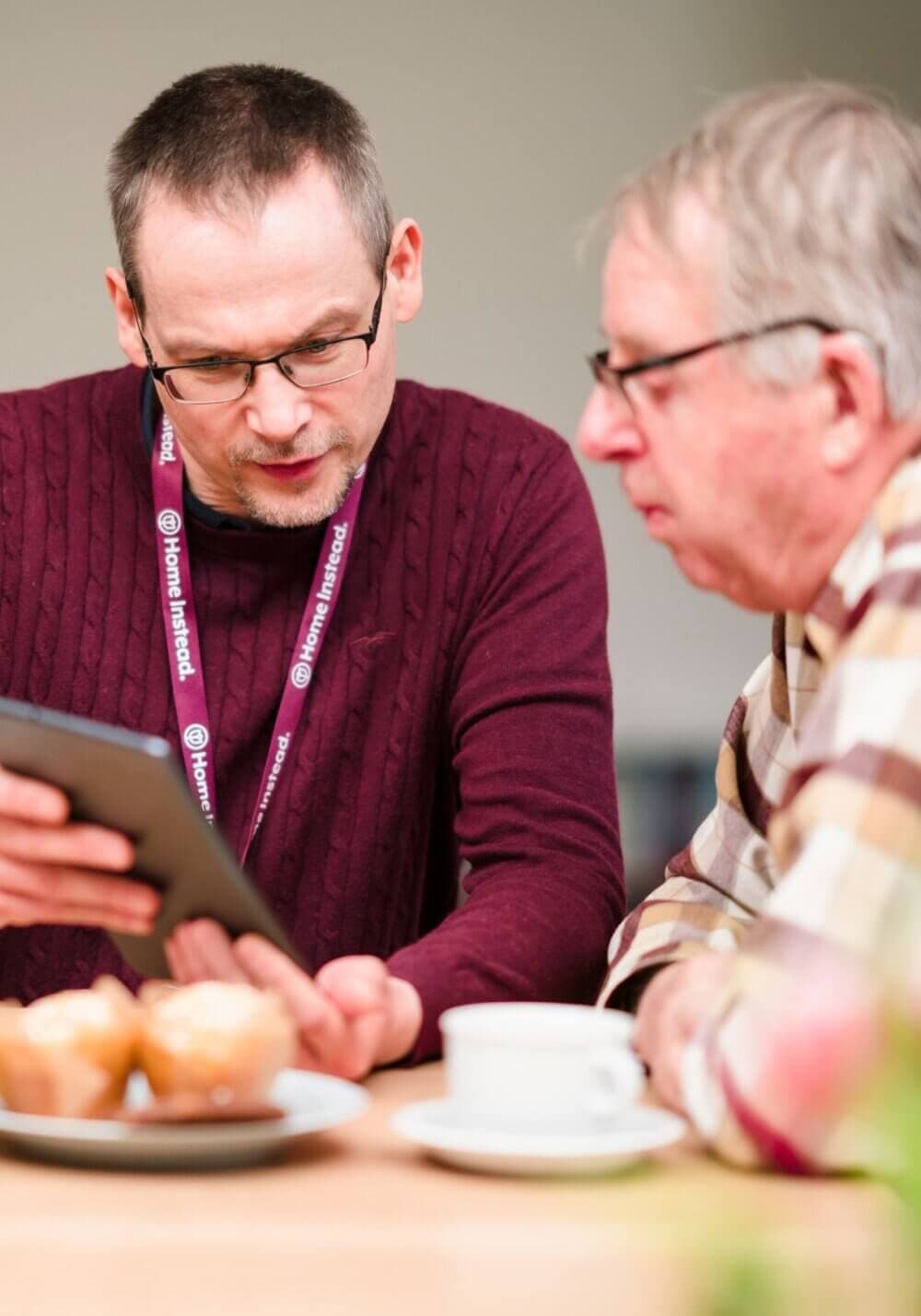 Two men are seated at a table with muffins and coffee, while one shows the other something on a tablet. - Home Instead