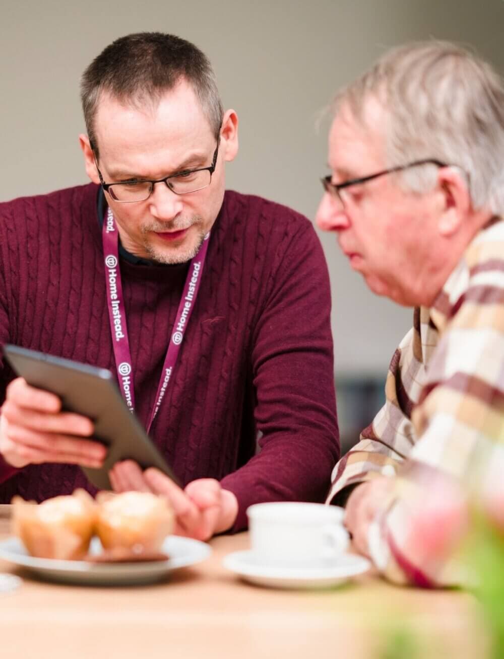 Two people sit at a table, one showing the other something on a tablet, with muffins and a cup in the foreground. - Home Instead