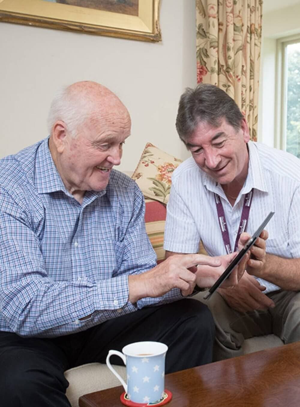 Two elderly men smiling and looking at a tablet while sitting on a couch with a coffee cup on the table. - Home Instead