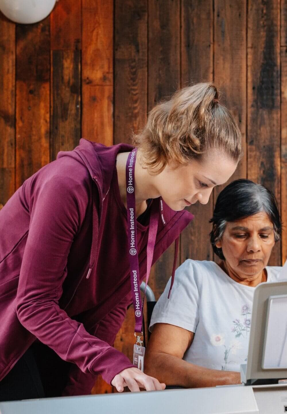 A caregiver helps an elderly woman who is using a laptop, both interacting in a wooden-floored room. - Home Instead