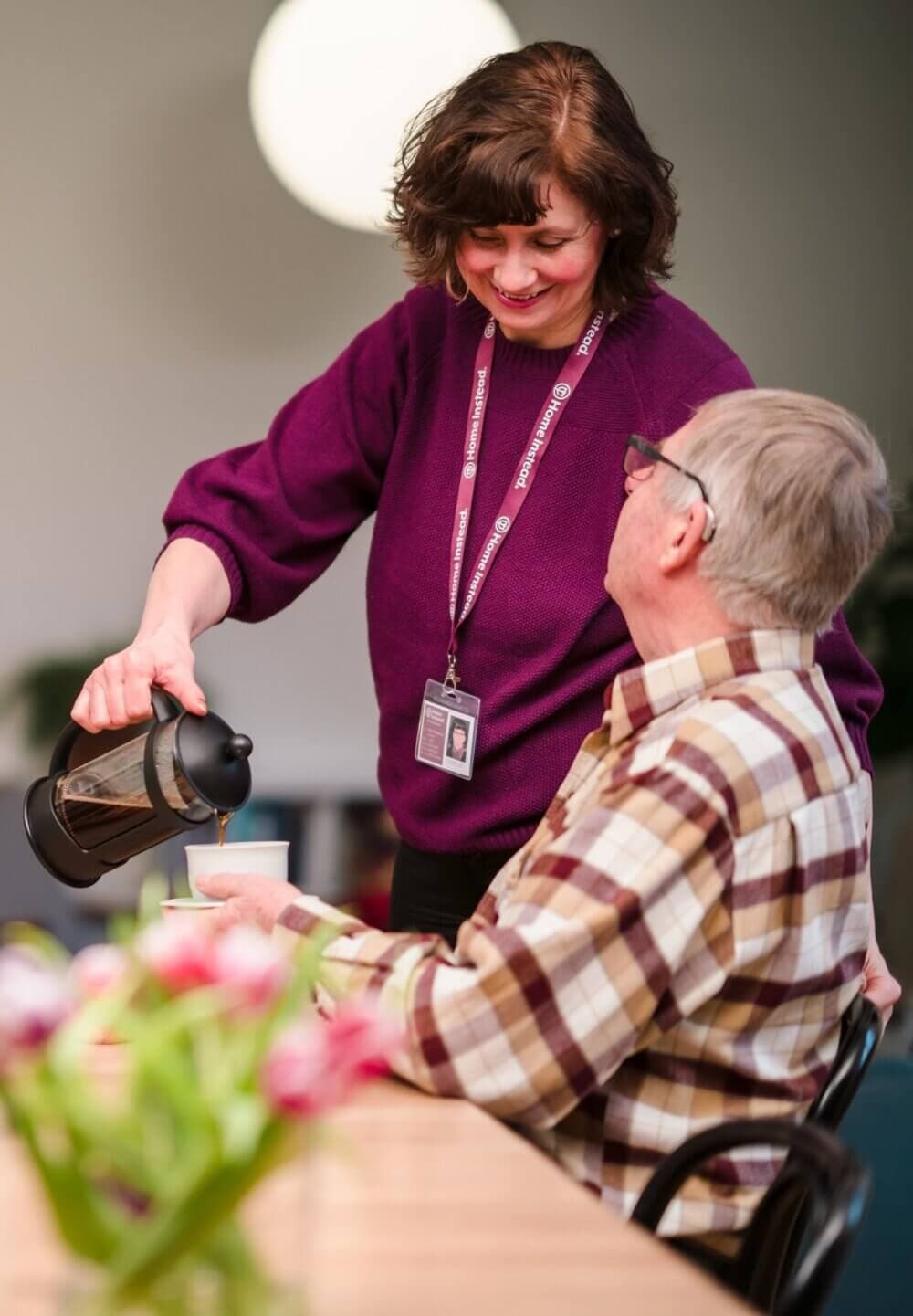 Woman in purple sweater pours coffee for seated man in plaid shirt at a table with flowers. - Home Instead