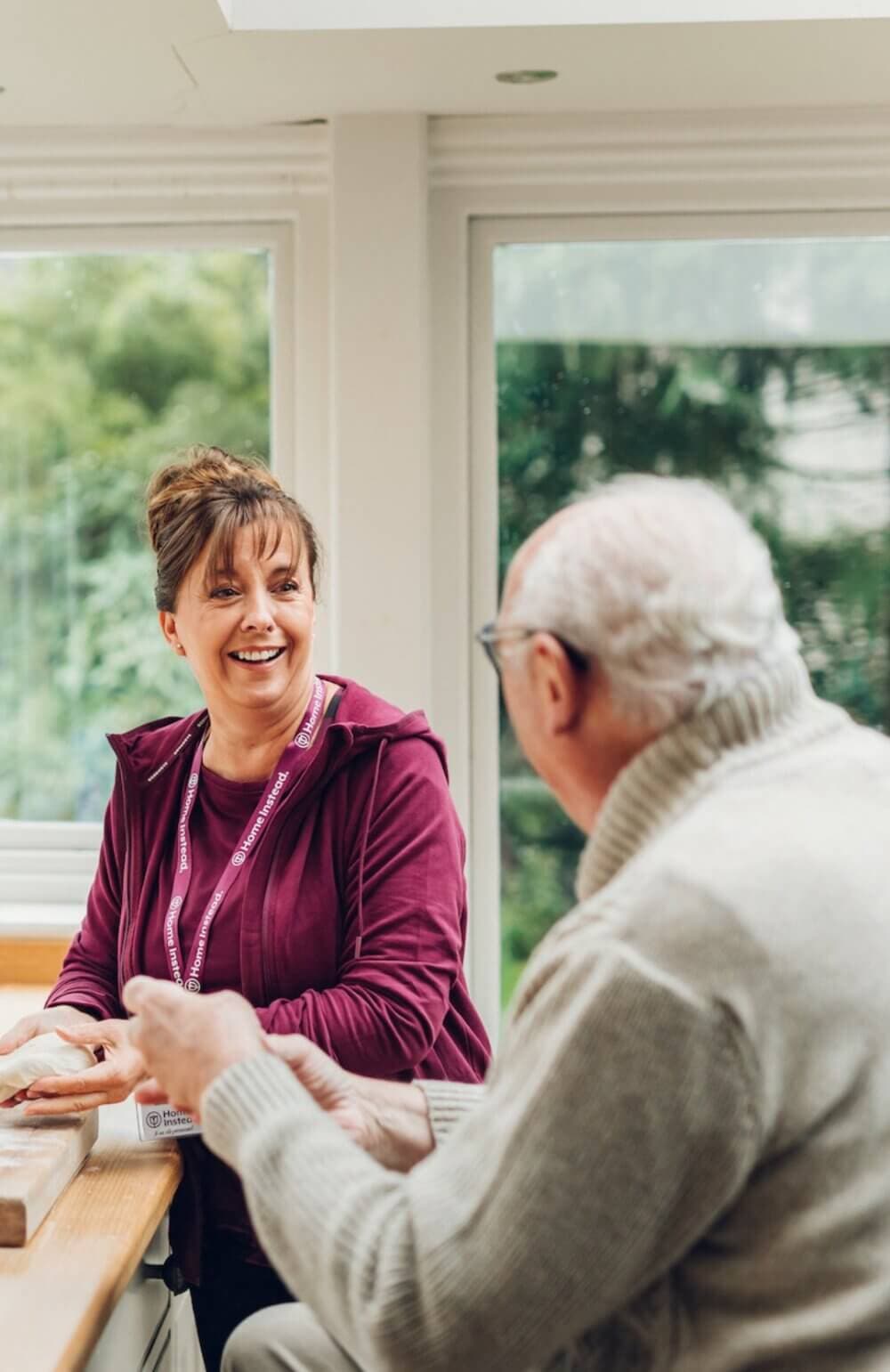A woman smiling and chatting with an elderly man in a light-filled room, both seated at a table. - Home Instead