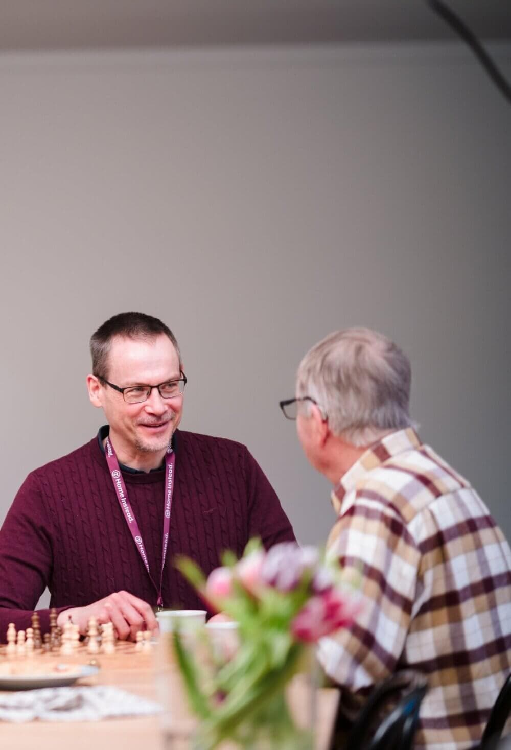 Two people engaged in conversation at a table with a chessboard, unclear pieces, and flowers in the foreground. - Home Instead