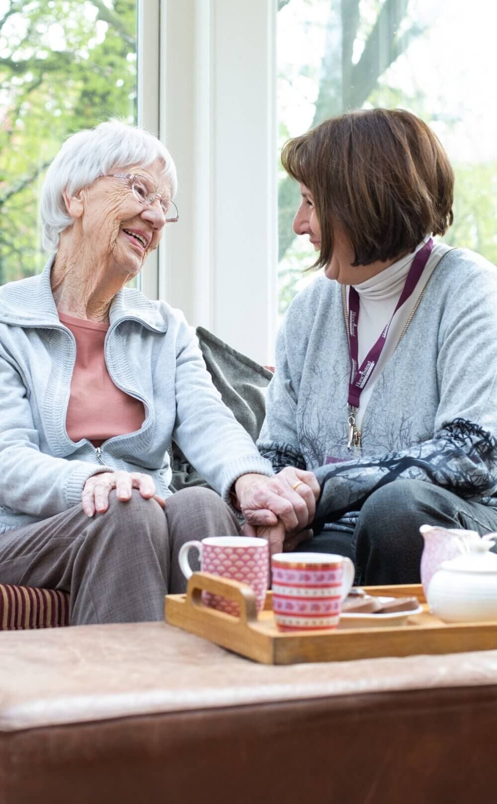 An elderly woman and a younger woman smiling at each other, sitting on a couch with tea set on a tray in front of them. - Home Instead