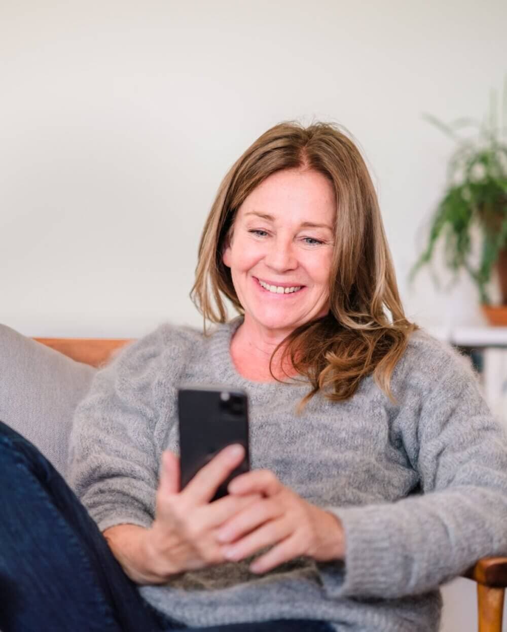 Woman smiling while looking at her phone, sitting on a couch with a plant in the background. - Home Instead