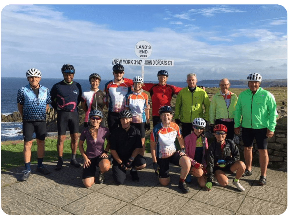 Group of cyclists in colorful gear posing in front of the Land's End sign with ocean and cliffs in the background. - Home Instead