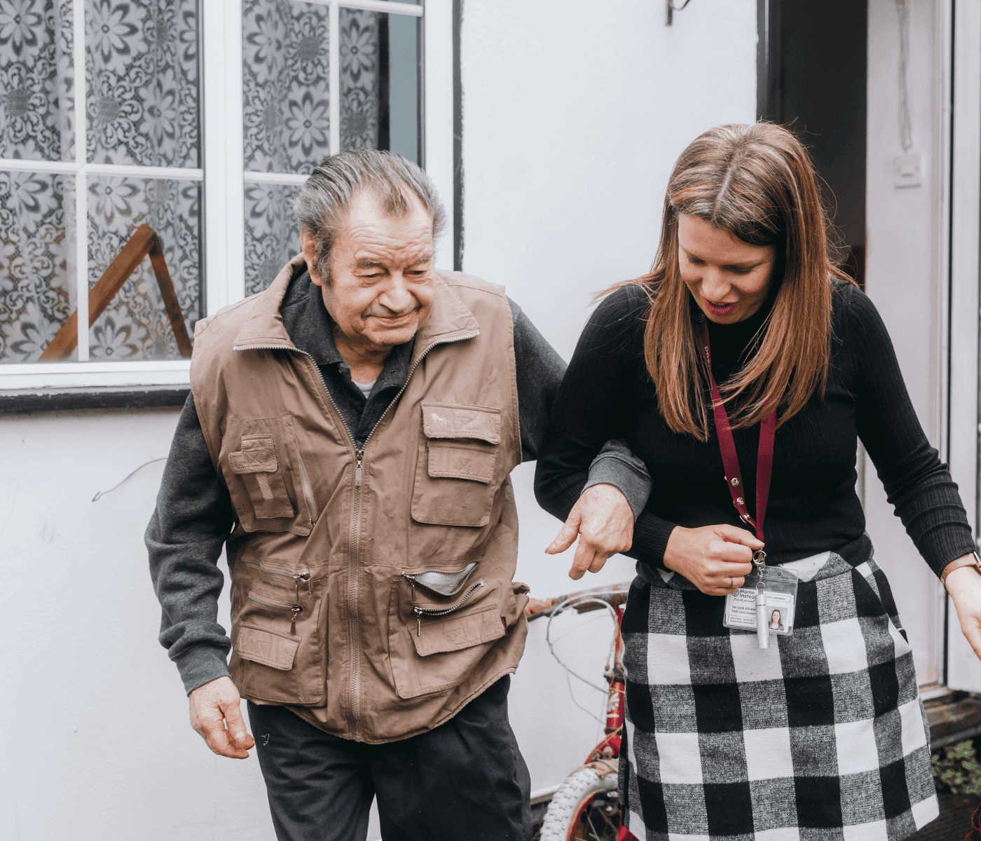 A woman assists an elderly man walking outside near a bicycle, with a patterned window and door in the background. - Home Instead