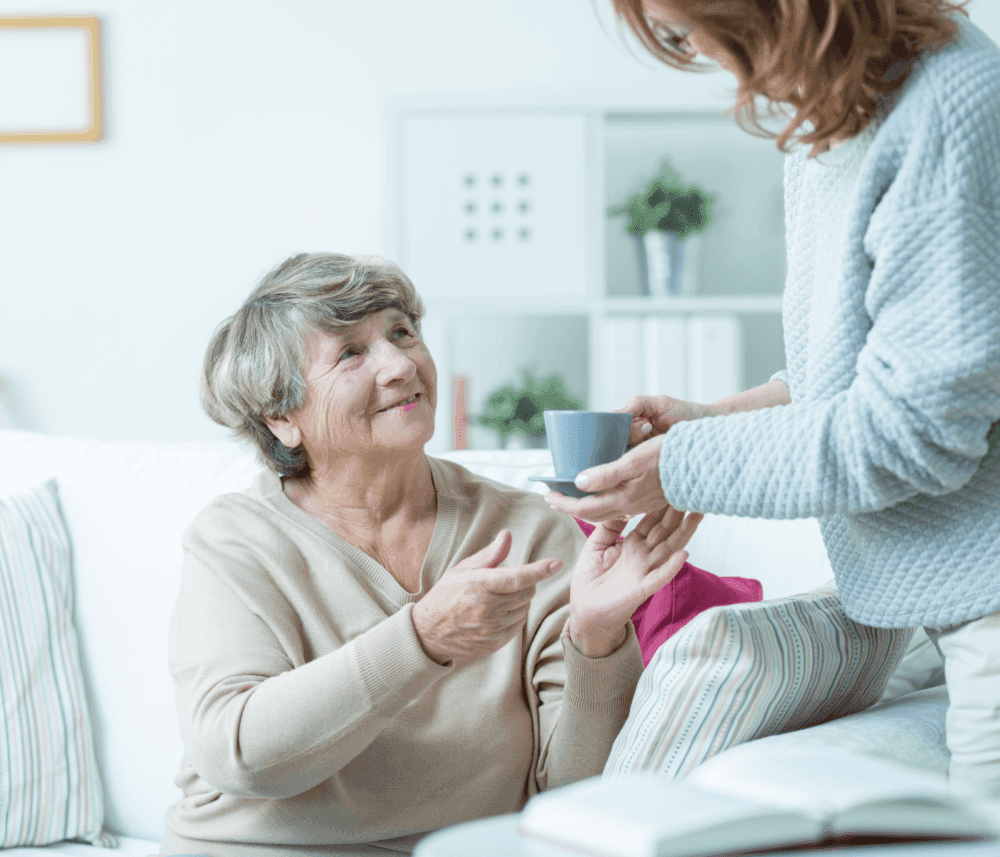 An elderly woman smiles while receiving a cup of tea from a younger person in a cozy living room. - Home Instead