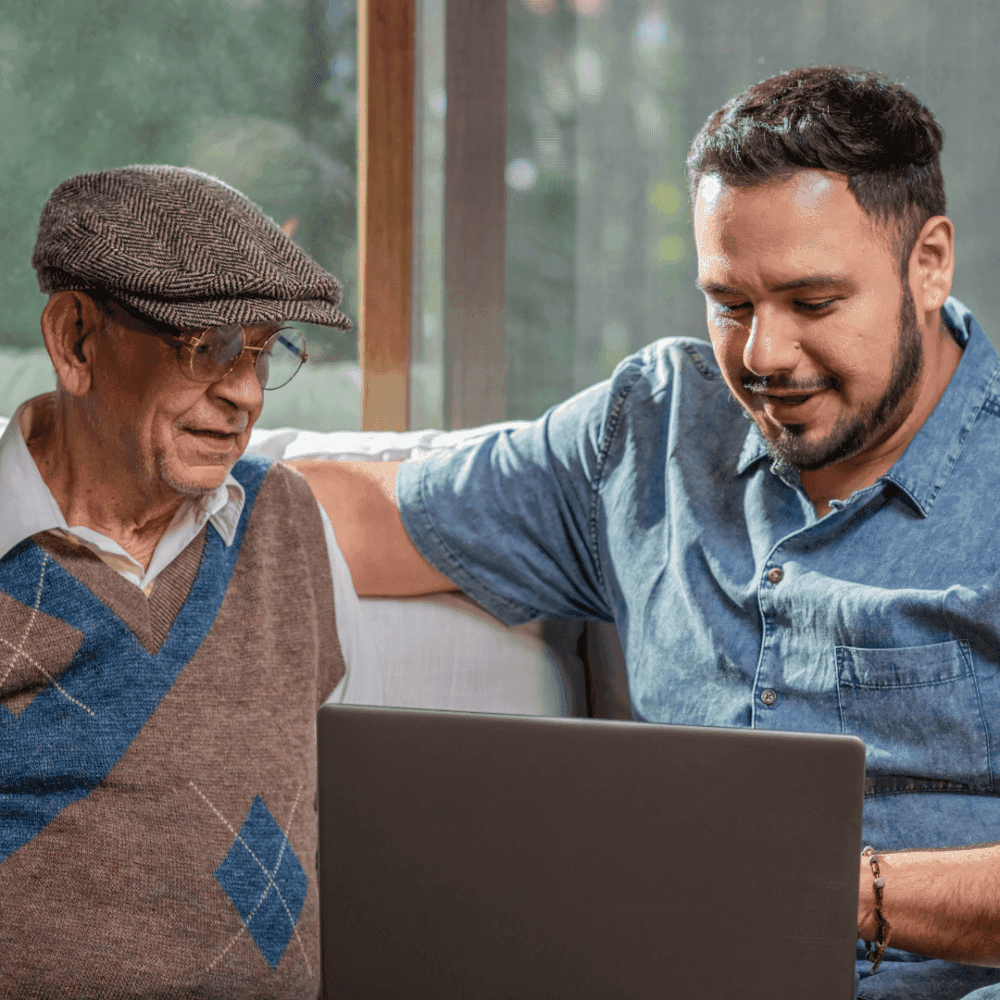 An elderly man and a younger man smiling while looking at a laptop screen, sitting on a couch together. - Home Instead