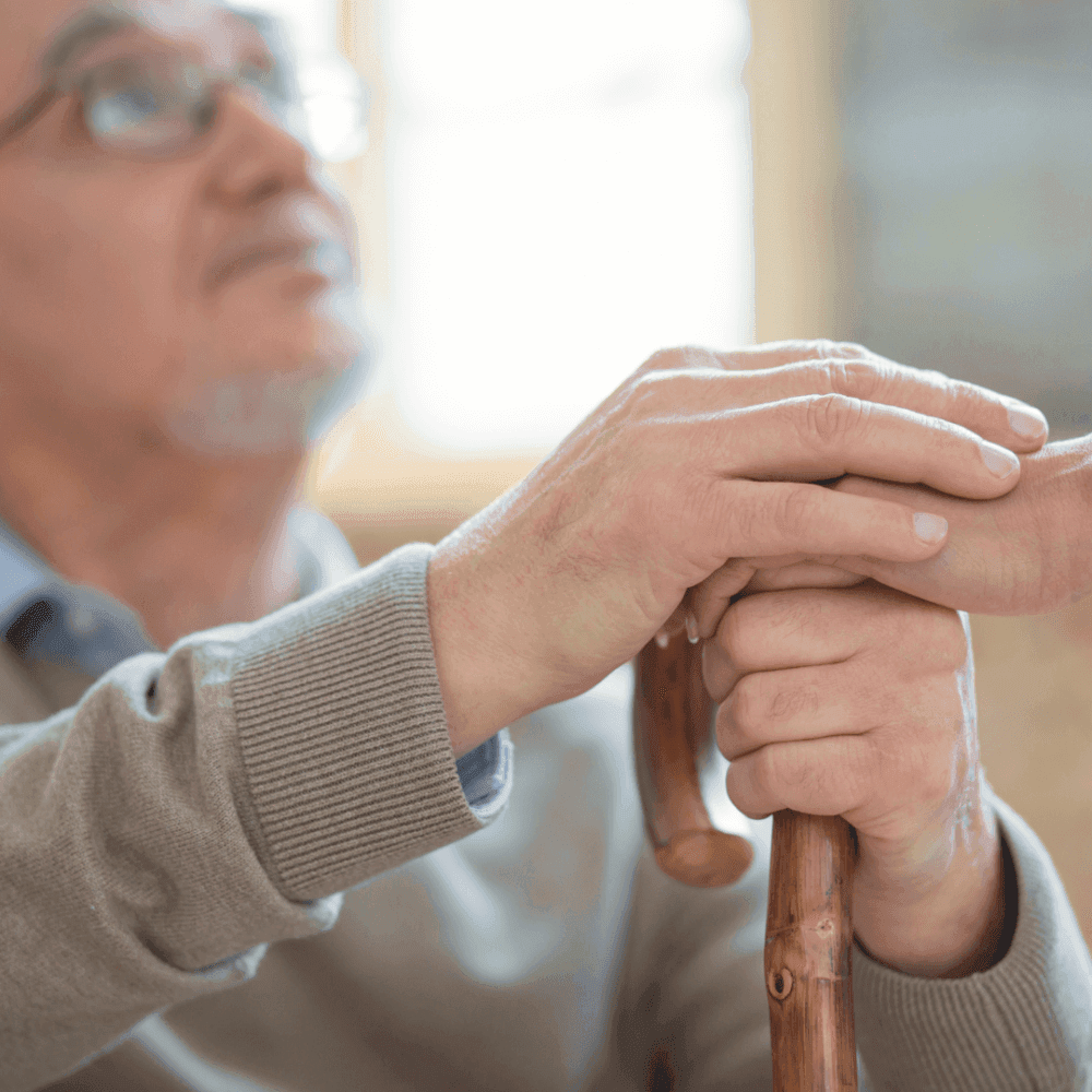 Older man holding a wooden cane with both hands, looking upward thoughtfully. - Home Instead