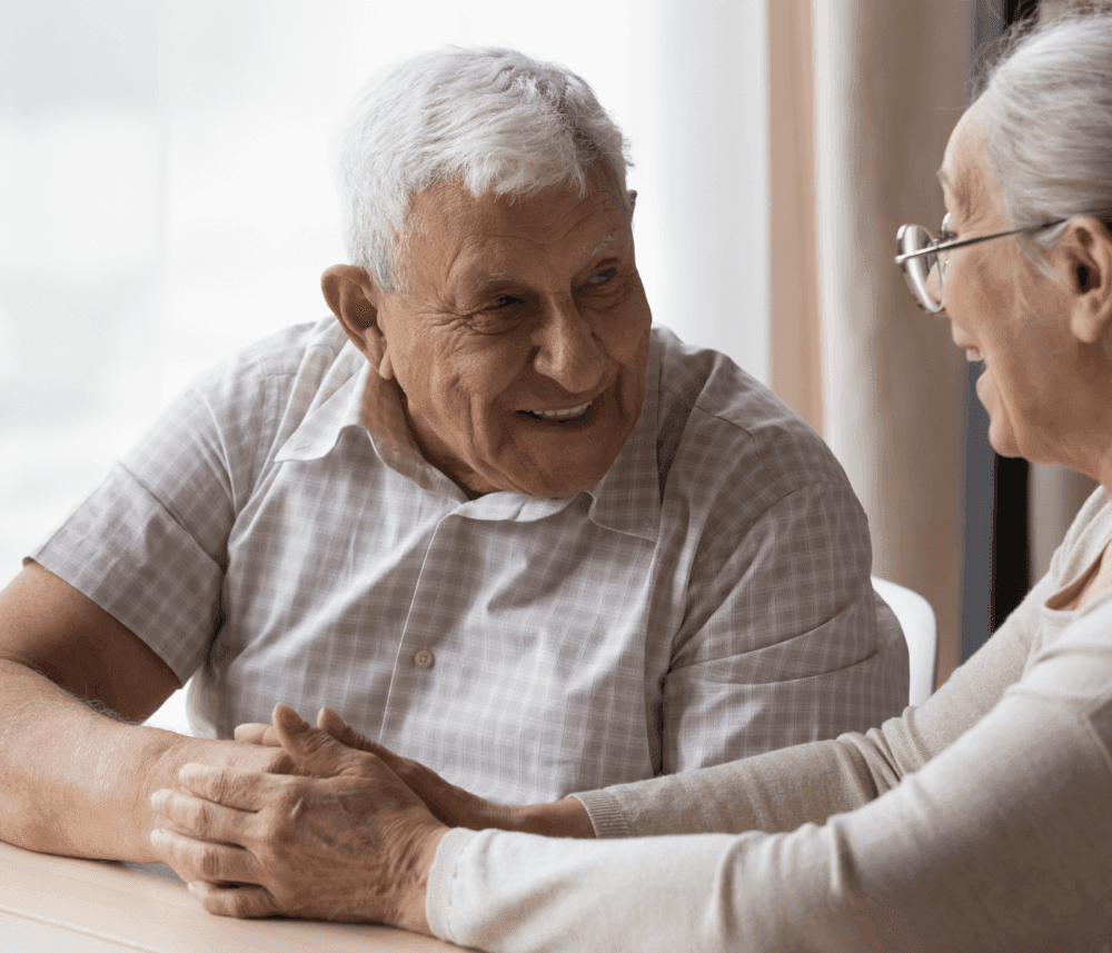 Elderly couple sitting at a table, holding hands and smiling at each other warmly. - Home Instead