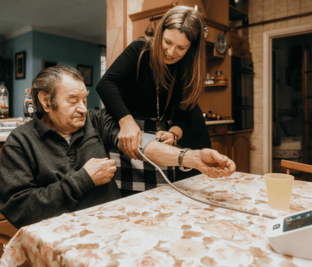 A woman helps an elderly man measure his blood pressure at home, both smiling and seated at a floral table. - Home Instead
