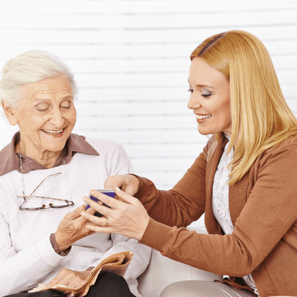 Young woman showing a smartphone to an elderly woman with a smile, the elderly woman holds a newspaper and her glasses. - Home Instead