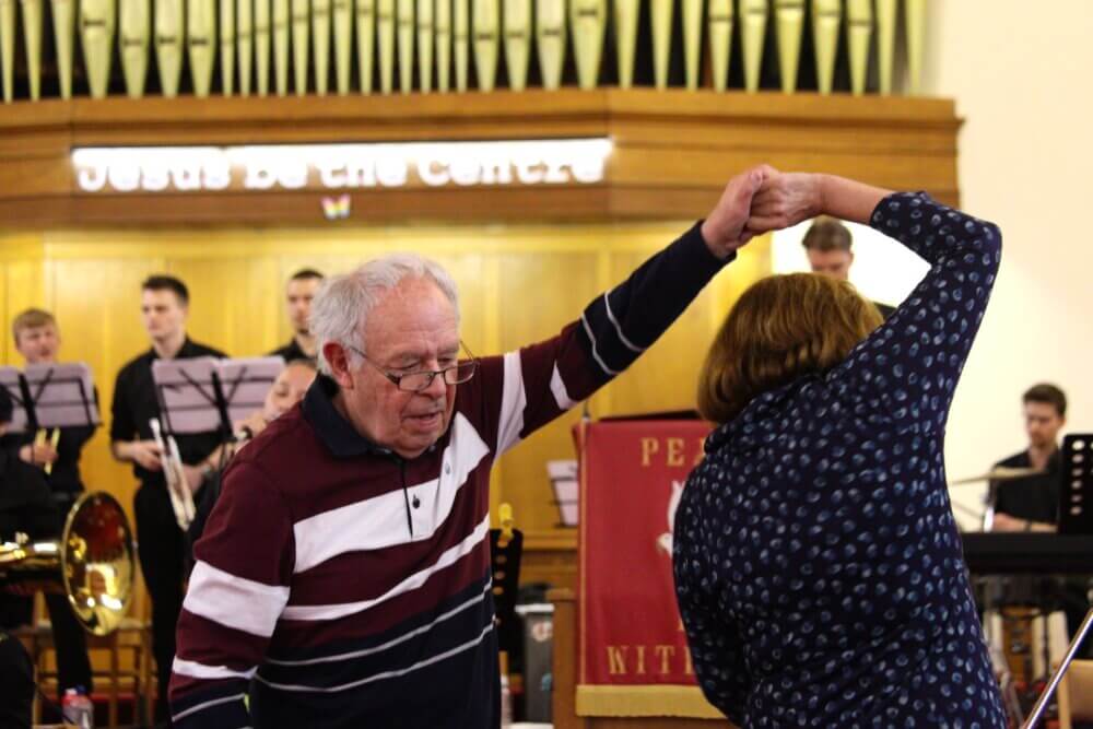 An elderly couple dances joyfully in front of a live band in a church hall. - Home Instead