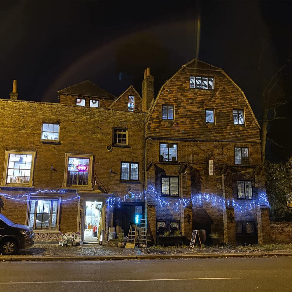 Brick building with lit windows and blue fairy lights hanging outside at night, with a neon sign and cars parked nearby. - Home Instead