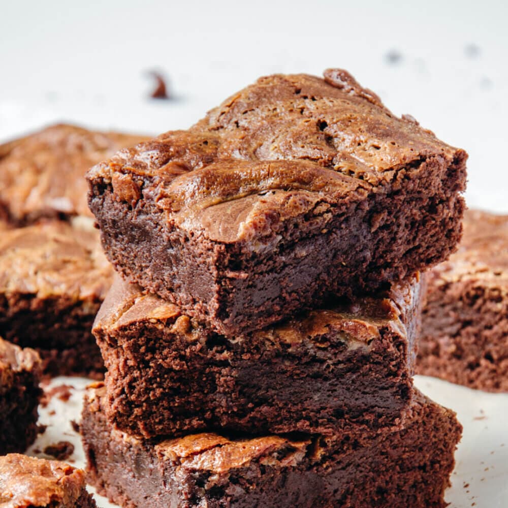 Close-up of a stack of three homemade chocolate brownies on a white surface, with more brownies in the background. - Home Instead