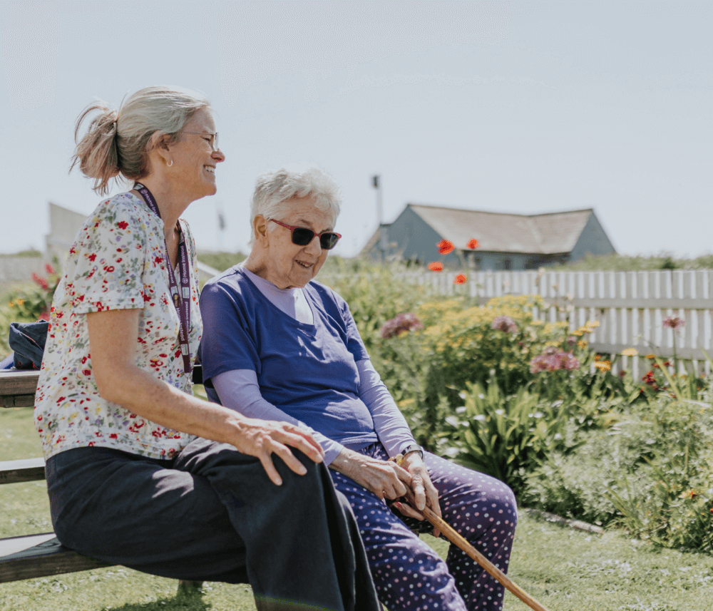 Two older women sitting on a bench outdoors, smiling and talking, with a garden and houses in the background. - Home Instead
