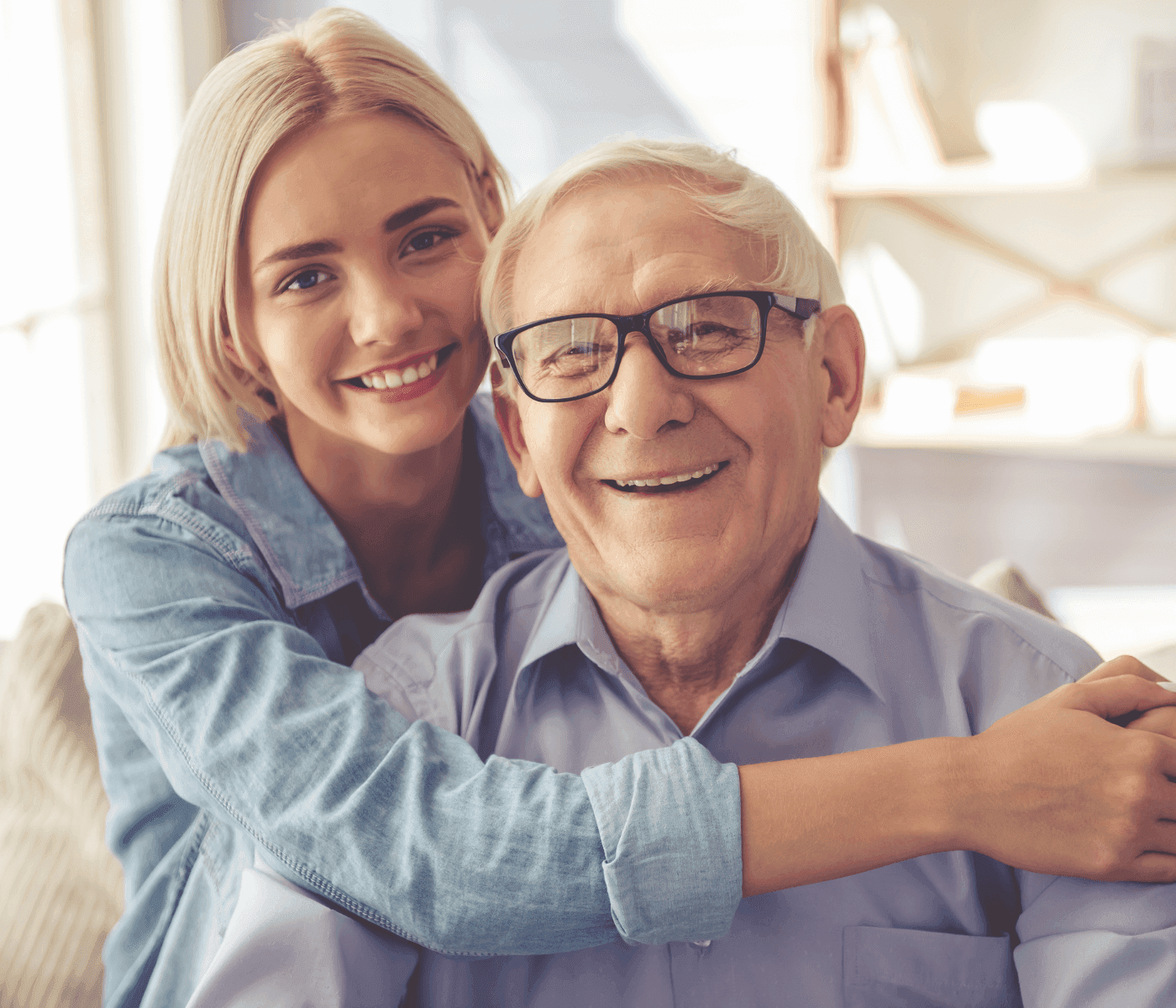 A young woman with blonde hair hugs an elderly man with glasses, both smiling warmly inside a cozy room. - Home Instead