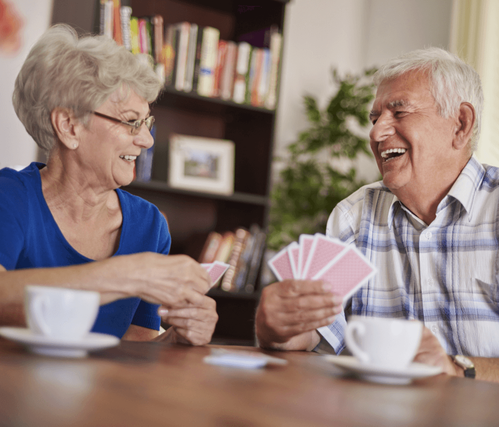 An elderly couple enjoys playing cards and laughing together at a table with coffee cups in a cozy room. - Home Instead