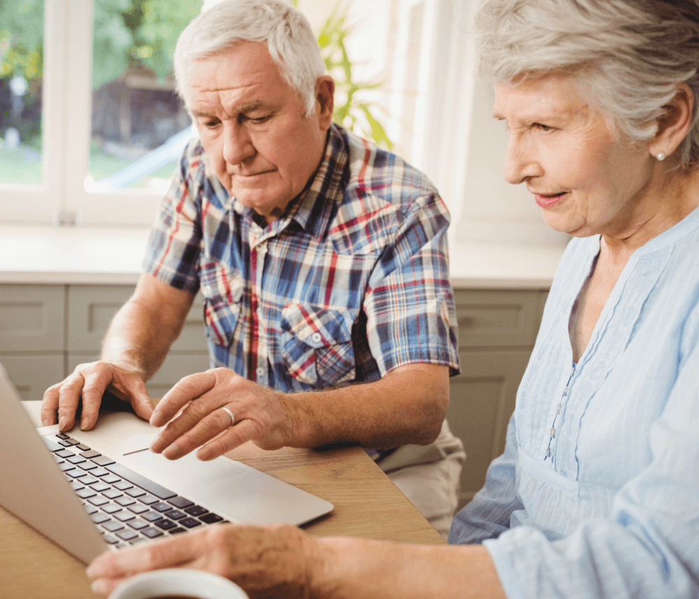 An elderly couple uses a laptop together at a wooden table in a bright room with large windows. - Home Instead