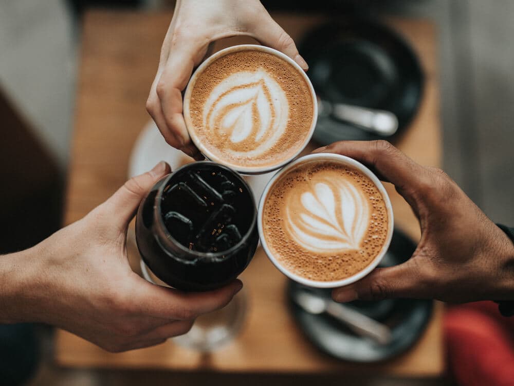 Three hands holding cups of coffee with latte art and a glass of iced coffee, over a wooden table. - Home Instead