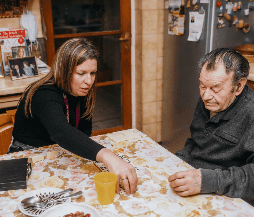 A woman is helping an elderly man play dominoes at a table in a kitchen. - Home Instead