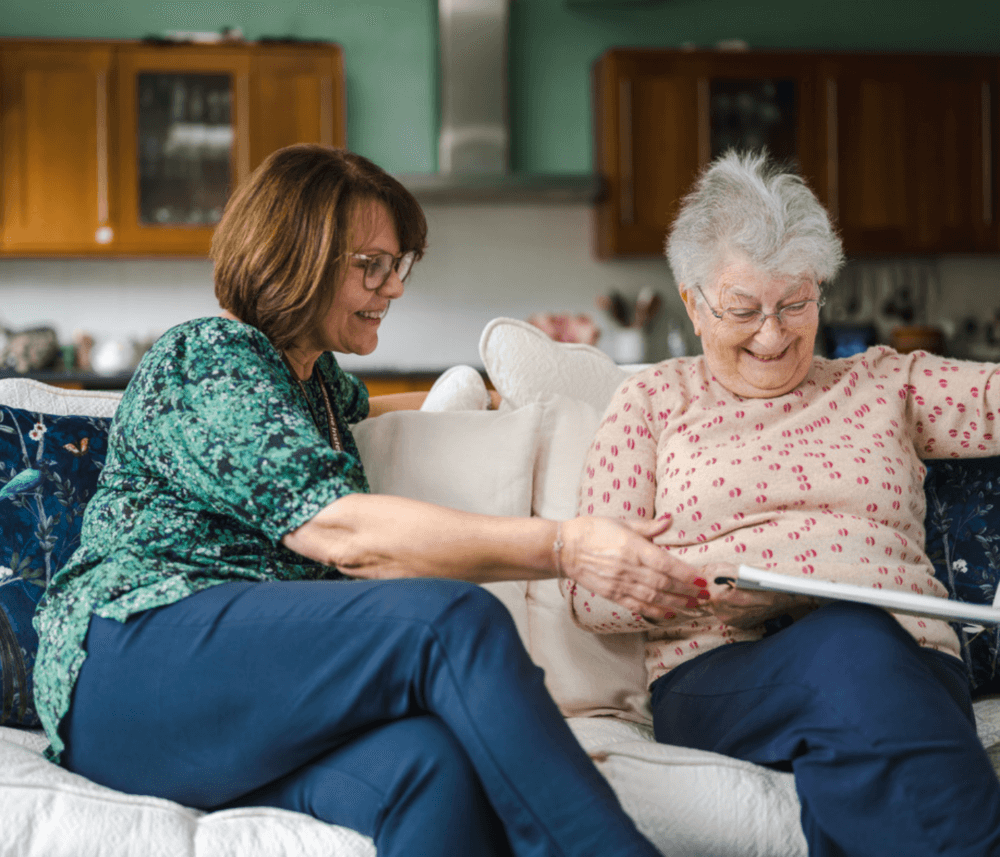 Two women sitting on a couch, one showing a tablet to the other; both smiling and engaged. - Home Instead