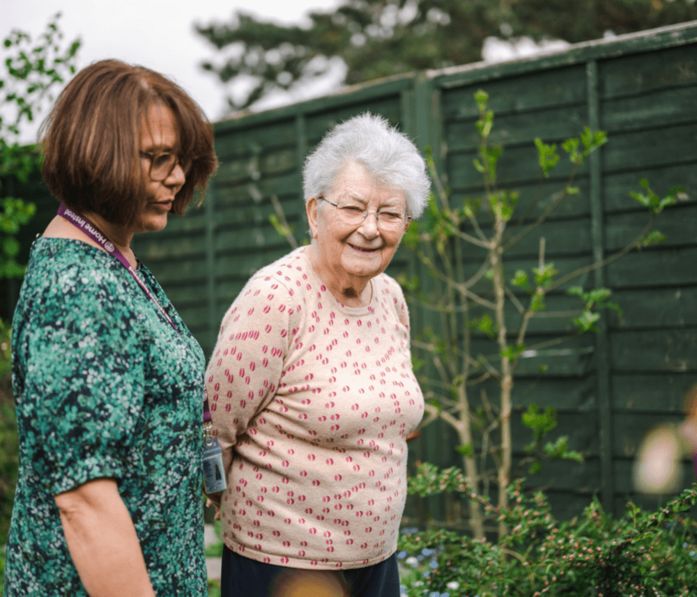 Elderly woman with short white hair and glasses smiling next to a younger woman in a garden. - Home Instead
