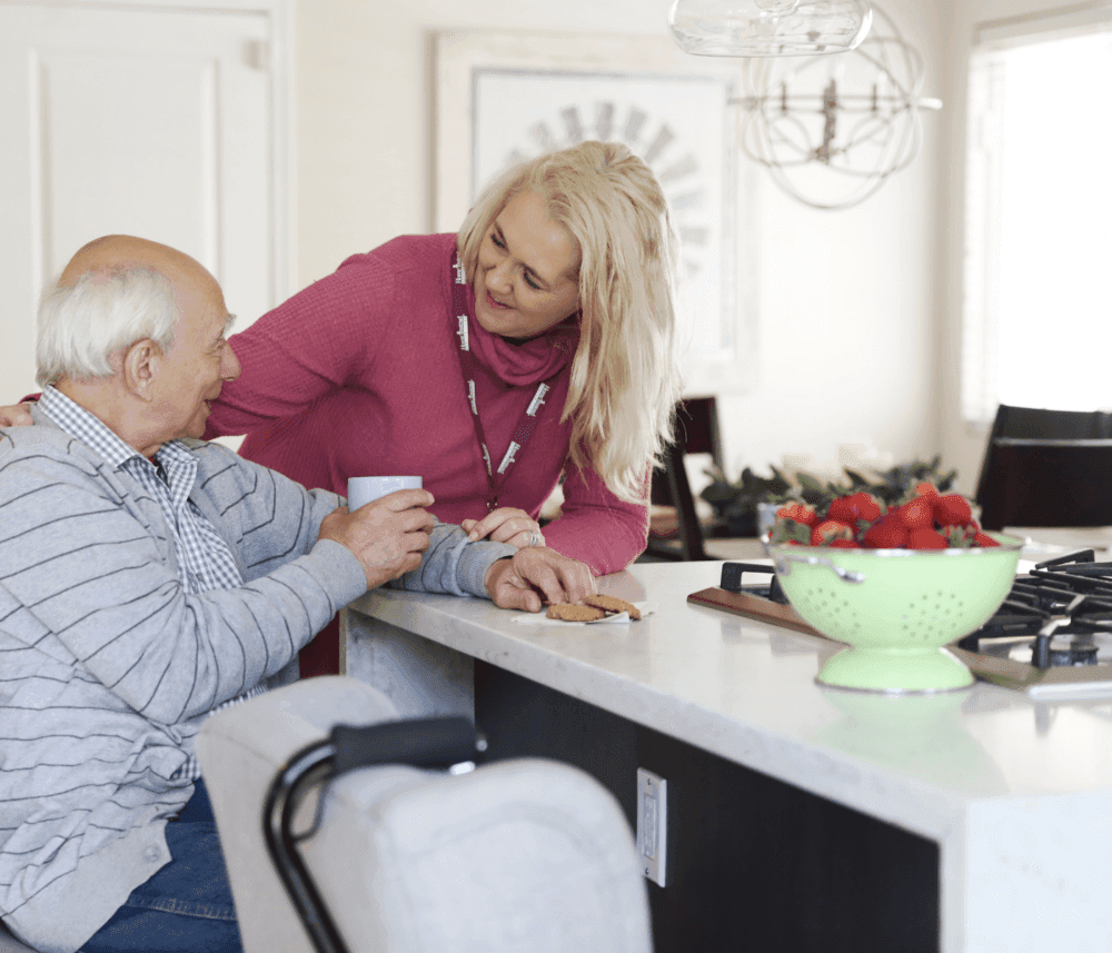 A woman in a pink sweater assists an elderly man with a drink in a kitchen with a bowl of strawberries. - Home Instead