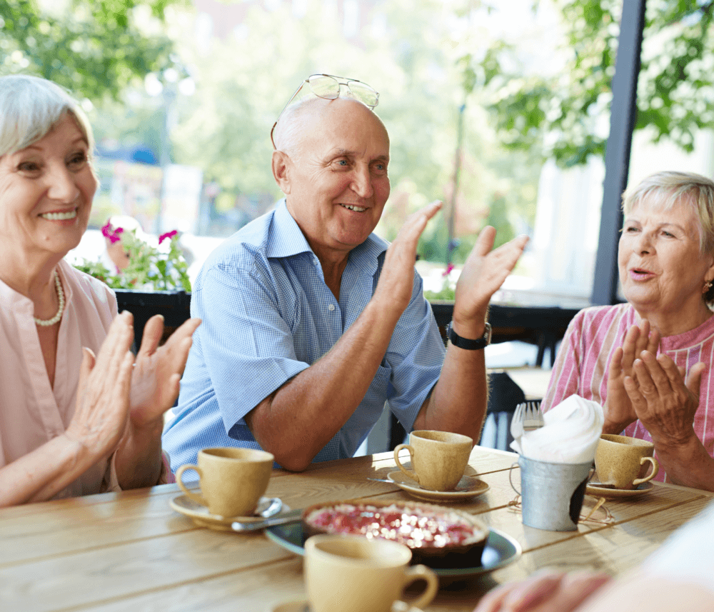 Three elderly people sitting at an outdoor café table, smiling and clapping, with coffee cups and pastries on the table. - Home Instead