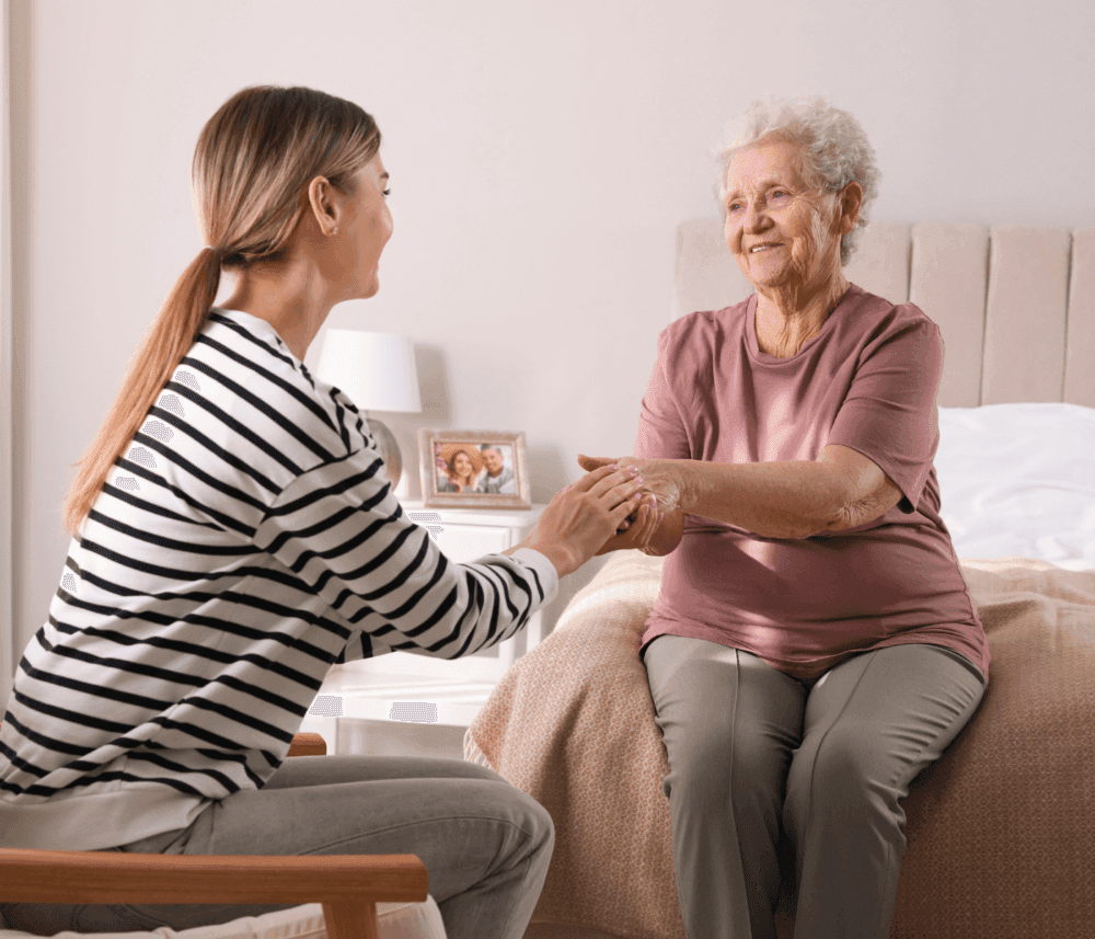 A young woman and an elderly woman are smiling and holding hands while sitting in a bedroom. - Home Instead