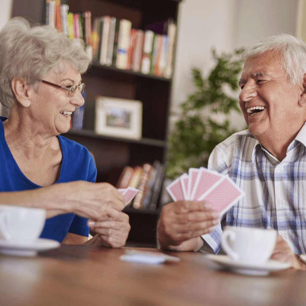 Two elderly people smiling and playing cards at a table with coffee cups in a cozy, book-filled room. - Home Instead